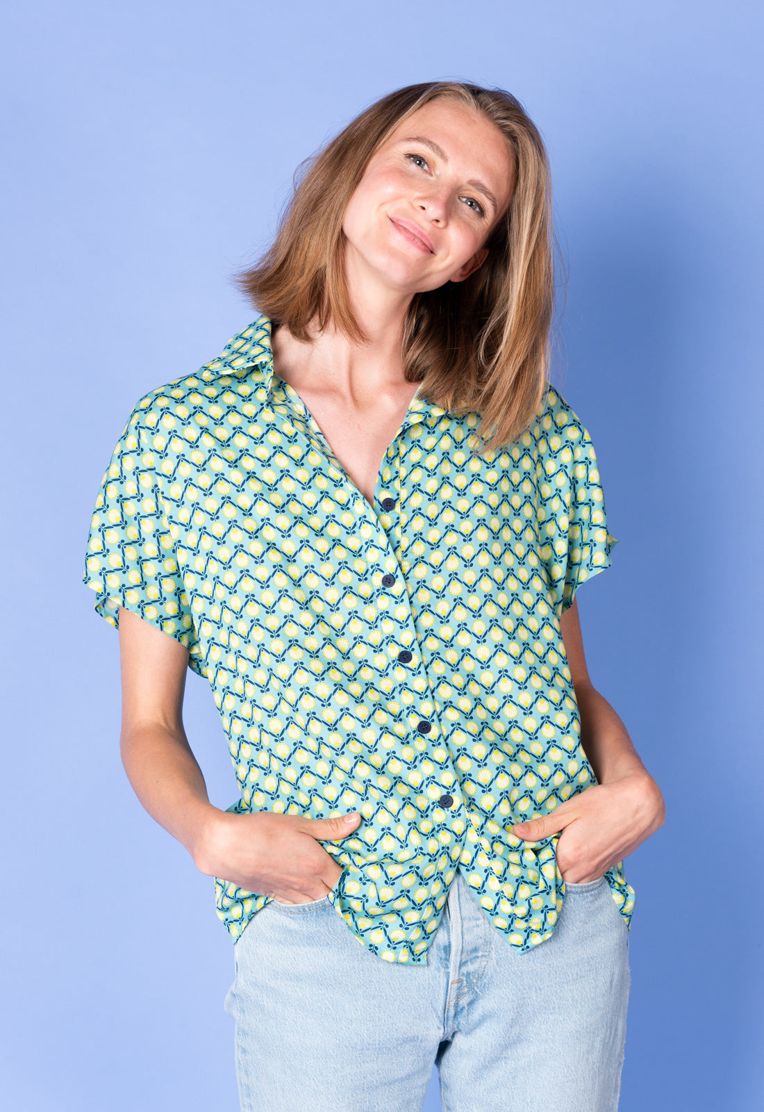 Lockeres Hemd - S25Top-R2 Das Bild zeigt eine Frau mit schulterlangem, leicht gewelltem, hellbraunem Haar. Sie trägt ein kurzärmeliges, locker geschnittenes Hemd mit einem auffälligen, ornamentalen Muster in Grün-, Gelb- und Blautönen. Das Hemd hat eine Knopfleiste mit dunklen Knöpfen und ist vorne locker geknotet oder leicht offen getragen. Sie kombiniert es mit einer hellblauen Jeans.
Die Frau hat eine freundliche, entspannte Ausstrahlung und lächelt leicht. Ihre Kopfhaltung ist leicht zur Seite geneigt.