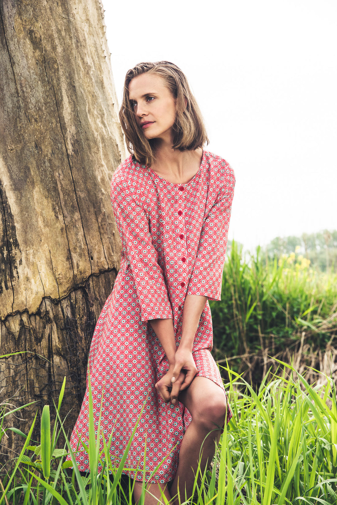  Langhaariges Model mit bunter Strickmütze, orange-dunkelblau gestreiften Strickpullover und beige-weinroter Hose vor einem Baum stehend. Moshiki